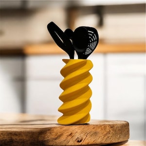 Peut inclure: Un pot à ustensiles jaune en forme de spirale avec des ustensiles de cuisine noirs. Le pot est en matériau solide et repose sur une surface en bois. Les ustensiles comprennent une cuillère ajourée, une spatule et une louche.