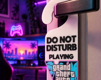 Op de afbeelding: Een witte deurhanger met zwarte tekst DO NOT DISTURB PLAYING en een kleurrijk Grand Theft Auto VI-logo met palmbomen, roze en blauwe accenten en een groot Romeins cijfer VI, hangend aan een zwarte metalen deurklink.