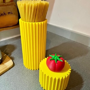 Può includere: Un alto contenitore cilindrico giallo brillante con scanalature verticali che contiene un mazzo di spaghetti secchi, accompagnato da un contenitore cilindrico giallo più corto abbinato con un coperchio a forma di pomodoro rosso e picciolo verde.