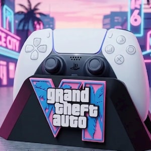 Könnte beinhalten: Ein weiß-schwarzer Videospiel-Controller steht in einem schwarzen Halter, der mit einer pink-blauen Grafikplatte und dem Schriftzug Grand Theft Auto VI versehen ist. Der Controller verfügt über Standardtasten, zwei Analogsticks und ein Touchpad.