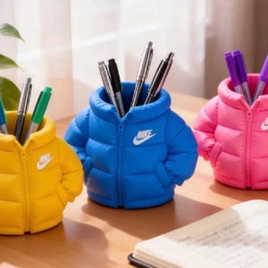 Peut inclure: Trois porte-stylos en forme de doudoune jaune, bleue et rose, arborant chacun un logo Nike blanc sur la poitrine. Les vestes présentent une texture matelassée, un détail de fermeture éclair centrale et contiennent divers stylos et crayons.