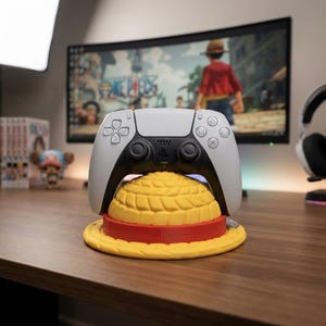 Pode incluir: Um comando de jogo sem fios branco e preto sobre um suporte em forma de chapéu de palha amarelo e vermelho. O suporte apresenta uma cúpula amarela texturizada com uma faixa vermelha e uma aba amarela larga para segurar o comando.