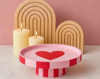 Puede incluir: Una bandeja redonda rosa con pedestal que presenta un gran corazón rojo en el centro y rayas verticales alternas rojas y rosas en la base y el borde, acompañada de dos velas de pilar color crema y dos paneles decorativos de madera clara.