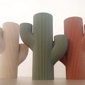 May include: Three cylindrical vases shaped like saguaro cacti, each featuring a vertical ribbed texture. The vases are colored in light beige, muted olive green, and terracotta orange, standing upright with two side arms extending from each main body.