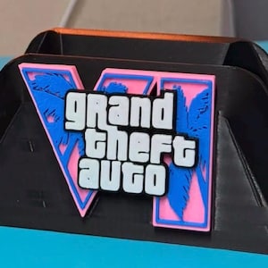 Puede incluir: Un soporte de pl&aacute;stico negro con el logotipo de Grand Theft Auto VI en la parte frontal. El logo consiste en un gran n&uacute;mero romano VI de color rosa con siluetas de palmeras azules, superpuesto con el texto blanco Grand Theft Auto en negrita.