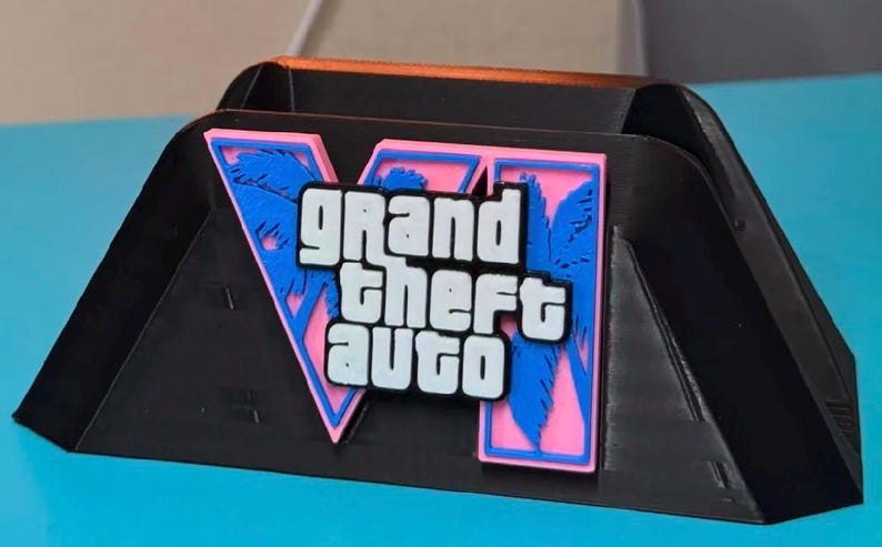 Puede incluir: Un soporte de pl&aacute;stico negro con el logotipo de Grand Theft Auto VI en la parte frontal. El logo consiste en un gran n&uacute;mero romano VI de color rosa con siluetas de palmeras azules, superpuesto con el texto blanco Grand Theft Auto en negrita.