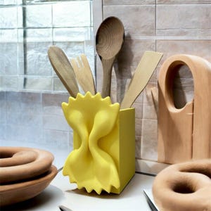 Peut inclure: Un porte-ustensiles jaune en forme de pâtes farfalle. Il contient des cuillères en bois, une fourchette et une spatule. L'ensemble est posé sur un plan de travail et un mur carrelé.