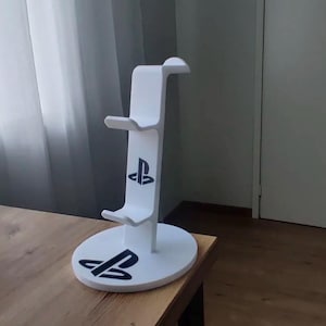 Puede incluir: Un soporte vertical blanco para mandos de juego con una base circular y dos ganchos escalonados para almacenamiento. El soporte está decorado con logotipos negros de PlayStation impresos en el frente de la columna vertical y en la base circular.