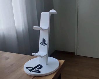 Op de afbeelding: Een witte verticale standaard voor gamecontrollers met een ronde voet en twee trapsgewijze haken voor opslag. De standaard is versierd met zwarte PlayStation-logo's die op de voorkant van de verticale steunkolom en op de ronde voet zijn gedrukt.