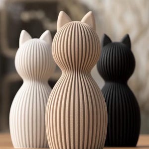 Puede incluir: Tres figuras con forma de gato con una textura acanalada vertical. La figura central es de color beige, mientras que las figuras laterales son blancas y negras. Cada pieza presenta una cabeza redondeada con orejas puntiagudas y un cuerpo cónico.