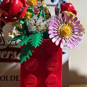 Może przedstawiać: Żywy bukiet kwiatów wykonany z klocków LEGO, w tym czerwone róże, żółte pompony i duża różowa stokrotka. Kwiaty ułożone są w czerwonym wazonie z klocków LEGO, z widocznym logo LEGO.