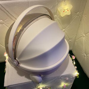 May include: A white, dome-shaped container with a clear top and a handle. The container sits on a white surface and is decorated with a string of lights with pink flowers and a smiling star-shaped light.