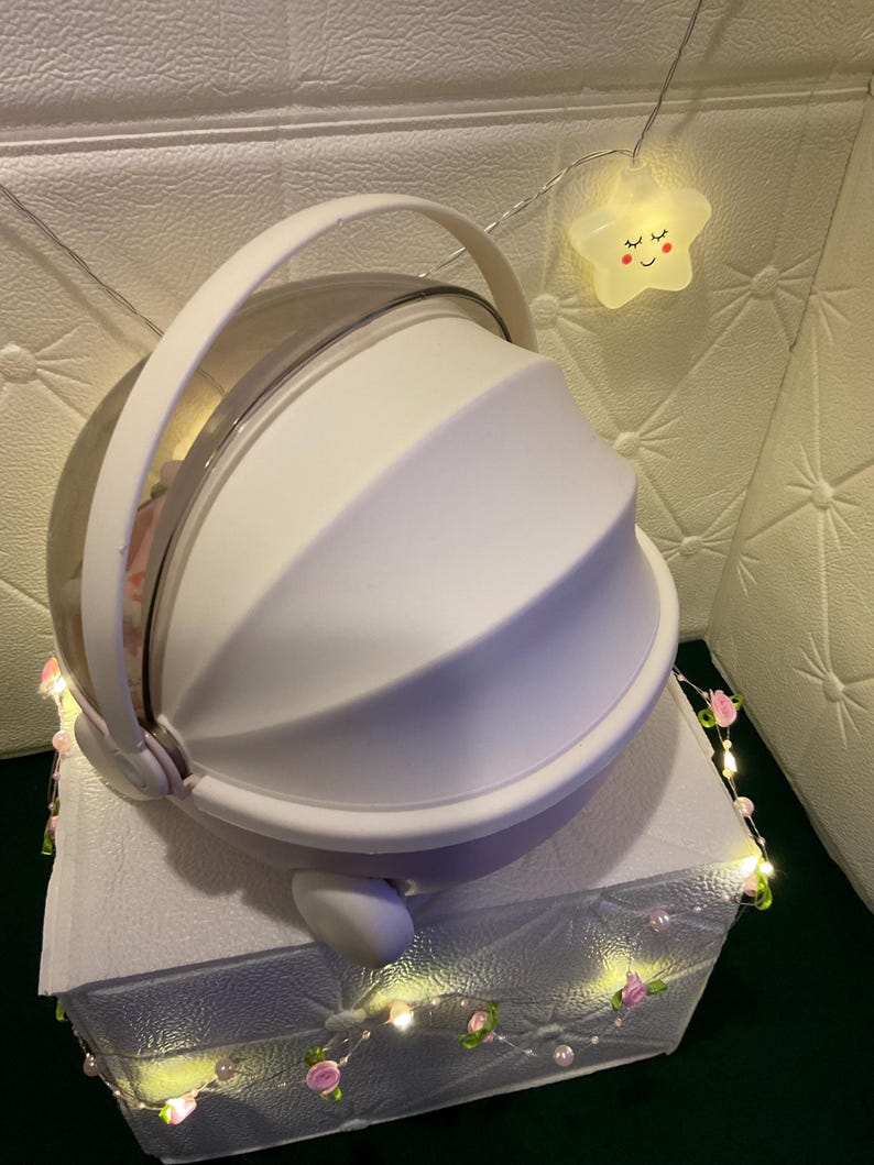 May include: A white, dome-shaped container with a clear top and a handle. The container sits on a white surface and is decorated with a string of lights with pink flowers and a smiling star-shaped light.