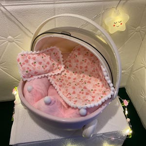 May include: A pink pet carrier with a clear dome and white handle. Inside, a pink plush bed, pillow, and blanket with a cherry pattern. The carrier sits on a white surface, decorated with fairy lights.