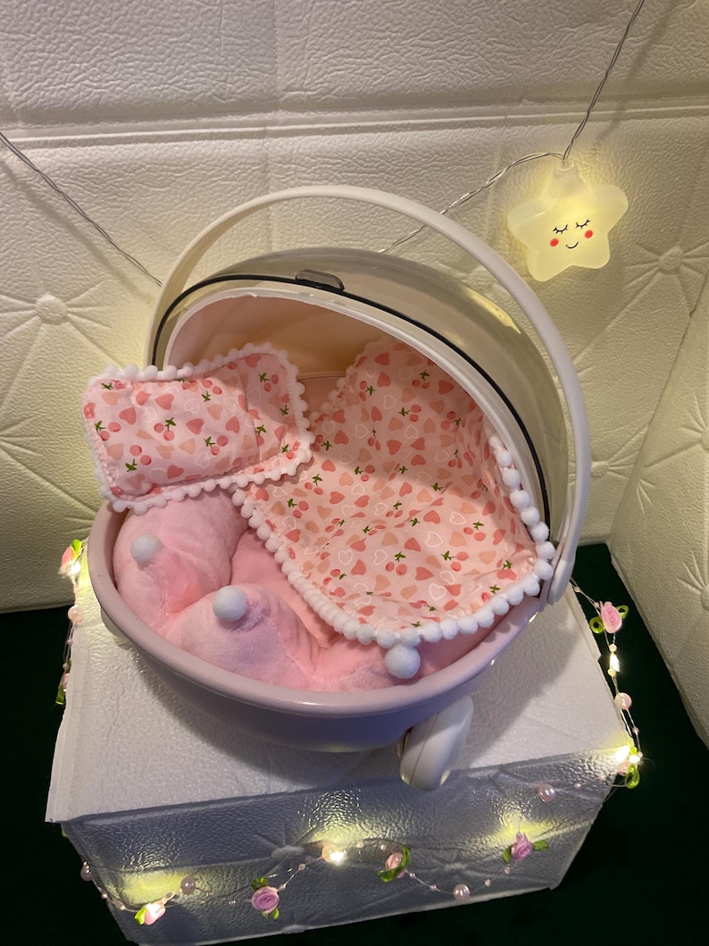 May include: A pink pet carrier with a clear dome and white handle. Inside, a pink plush bed, pillow, and blanket with a cherry pattern. The carrier sits on a white surface, decorated with fairy lights.