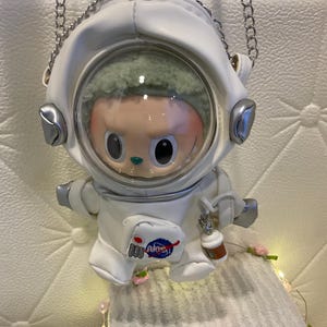 May include: A white astronaut-shaped crossbody bag featuring a clear circular visor revealing a doll with green hair inside. The bag has silver metallic accents, a silver chain strap, and a small patch on the front with red, white, and blue details.
