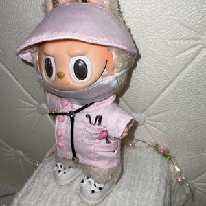 May include: A plush rabbit doll with large eyes, wearing a light pink visor, a matching pink medical-style jacket with buttons, a silver face mask, and a black stethoscope around its neck. The doll wears white shoes with black polka dots.