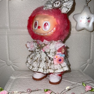 May include: A doll with a pink furry head and a white and gold dress. The doll has a bow on its head and is holding a pink camera. The doll is wearing white shoes and is standing on a white surface.