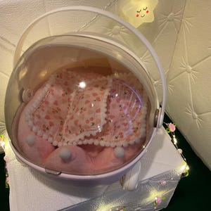 May include: A pink pet carrier with a clear, rounded top and a white handle. Inside, a pink bed with a cherry pattern and white trim is visible. The carrier sits on a white surface, with decorative lights and a star-shaped light in the background.