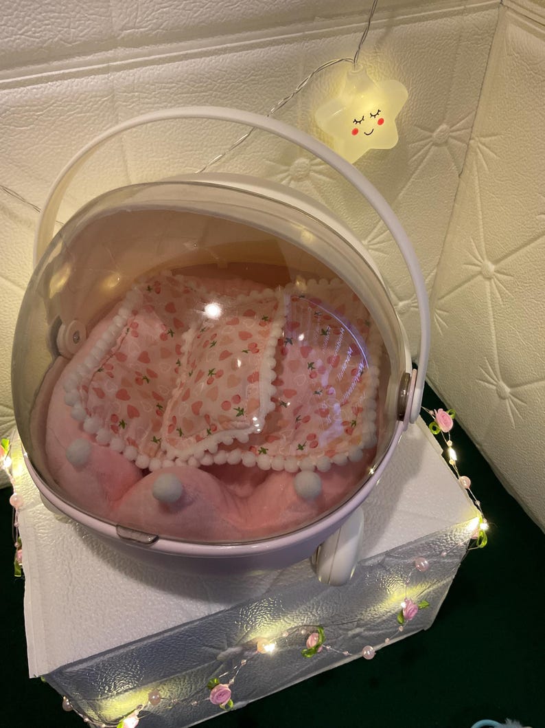 May include: A pink pet carrier with a clear, rounded top and a white handle. Inside, a pink bed with a cherry pattern and white trim is visible. The carrier sits on a white surface, with decorative lights and a star-shaped light in the background.