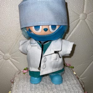 May include: A plush doll dressed as a medical professional, wearing a light blue surgical cap, a matching blue face mask, a white lab coat with buttons, a teal scrub top, and teal pants. A black stethoscope hangs around the neck of the doll.