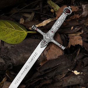 May include: A detailed image of a decorative sword with an ornate silver handle and a blade inscribed with text. The handle features red gemstone accents. The sword rests on a bed of dried leaves and wood.