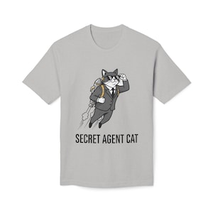 以下が含まれることがあります： スーツとサングラスをかけた猫の漫画が描かれたライトグレーのTシャツ。ジェットパックを背負い、「SECRET AGENT CAT」の文字がプリントされています。猫は空中を飛行しており、煙の跡が描かれています。