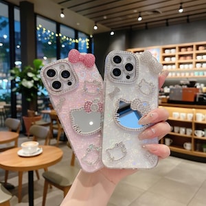 Puede incluir: Dos fundas de tel&eacute;fono con un dise&ntilde;o de Hello Kitty. Una es rosa con un lazo rosa, la otra blanca con un lazo plateado. Ambas fundas tienen un espejo y est&aacute;n decoradas con pedrer&iacute;a.