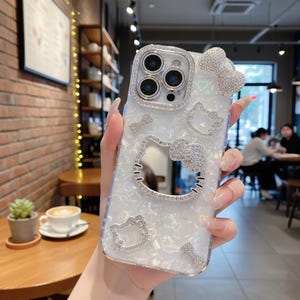 Puede incluir: Funda transparente para tel&eacute;fono con acabado nacarado, adornada con brillantes pedrer&iacute;a. El dise&ntilde;o presenta un lazo, corazones y m&uacute;ltiples contornos del personaje Hello Kitty. La lente de la c&aacute;mara est&aacute; rodeada por un anillo de pedrer&iacute;a.