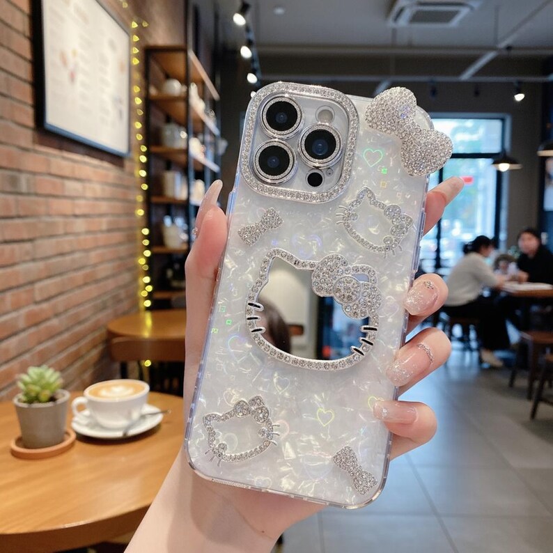 Puede incluir: Funda transparente para tel&eacute;fono con acabado nacarado, adornada con brillantes pedrer&iacute;a. El dise&ntilde;o presenta un lazo, corazones y m&uacute;ltiples contornos del personaje Hello Kitty. La lente de la c&aacute;mara est&aacute; rodeada por un anillo de pedrer&iacute;a.