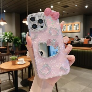 Puede incluir: Funda de tel&eacute;fono rosa con dise&ntilde;o de Hello Kitty. La funda presenta un lazo brillante, un espejo y detalles de pedrer&iacute;a. La lente de la c&aacute;mara est&aacute; rodeada por un borde plateado. La funda tiene un acabado nacarado.