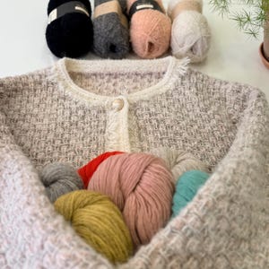 May include: A light-colored tweed jacket with a cream-colored trim and a single gold button. Several skeins of yarn in various colors, including black, gray, pink, and white, are arranged above the jacket. Additional yarn in various colors is inside the jacket.