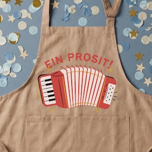 May include: A tan apron with a red accordion graphic and the words "EIN PROSIT!". The apron is on a blue background scattered with gold, white, and blue confetti. The text "the confetti department" is at the top.