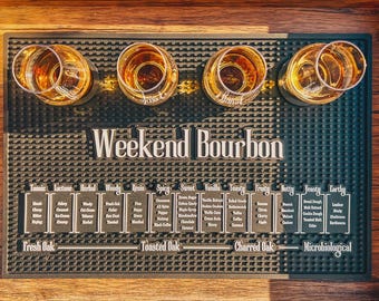 May include: A black rubber bar mat with the text "Weekend Bourbon" and four shot glasses filled with amber liquid. Below the text are flavor profiles such as "Fresh Oak", "Toasted Oak", and "Charred Oak".