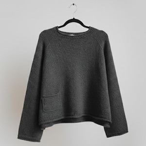 Puede incluir: Un suéter gris oscuro de gran tamaño con cuello redondo y un solo bolsillo en la parte delantera. El suéter está colgado de una percha negra sobre un fondo blanco liso. La prenda de punto parece ser un material suave y texturizado.