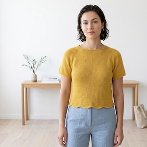 Puede incluir: Top de punto amarillo mostaza de manga corta con dobladillo festoneado. El top tiene un cuello redondo y se combina con pantalones azul claro. Una mesa de madera y una bolsa de lona están en el fondo.