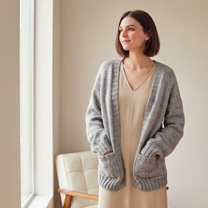 May include: A woman wearing a grey, open-front cardigan with two front pockets over a beige dress. The cardigan is a chunky knit, and the dress has a V-neck. The woman is standing near a window, with a white chair visible in the background.