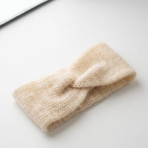 May include: A beige knitted headband with a twisted knot design. The headband is made of a soft, textured material and is designed to be worn around the head. The neutral color makes it a versatile accessory.