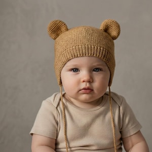 Puede incluir: Un gorro de bebé tejido de color beige con orejas de oso. El gorro tiene solapas para las orejas con cuerdas adjuntas. El bebé lleva un body beige. El fondo es de color neutro.
