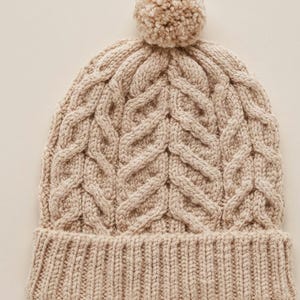 May include: A beige cable-knit beanie with a ribbed brim and a fluffy pom-pom on top. The hat features a textured, woven pattern throughout the crown. This warm winter hat is perfect for cold weather.