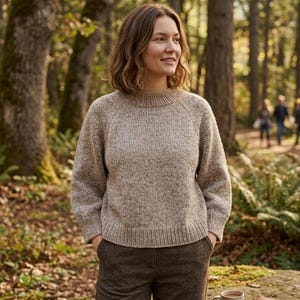 May include: A woman wearing a light brown, crew-neck sweater and brown trousers stands in a forest. The sweater has a textured knit pattern. A mug of coffee and a small notebook rest on a rock nearby.