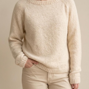 May include: Cream-colored knit sweater with a crew neck and raglan sleeves. The sweater has a textured appearance and ribbed cuffs and hem. The model is wearing beige pants. The overall style is casual and comfortable.
