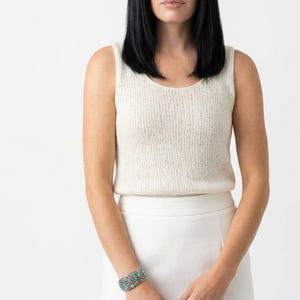 Puede incluir: Una mujer con un top sin mangas color crema y una falda blanca. También lleva una pulsera plateada con detalles turquesas. La mujer tiene el pelo negro.