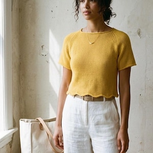 Puede incluir: Una mujer con un top de punto amarillo mostaza con dobladillo festoneado y pantalones de lino blancos. Un bolso tote beige con asas de cuero está sobre un banco de madera. El fondo es una pared de tonos neutros.