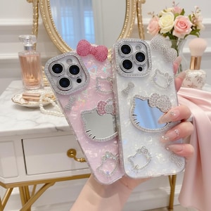 May include: Two decorative phone cases, one pink and one white, featuring a Hello Kitty design with a mirror face. Both cases are embellished with sparkling rhinestones and bows. The cases are held up in front of a vanity mirror and a bouquet of flowers.