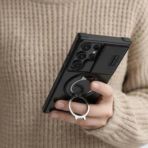 May include: Black phone case with a camera cover and a silver ring holder. The case has a matte finish and a raised edge around the camera lenses. The ring holder is attached to the back of the case and can be used as a stand.