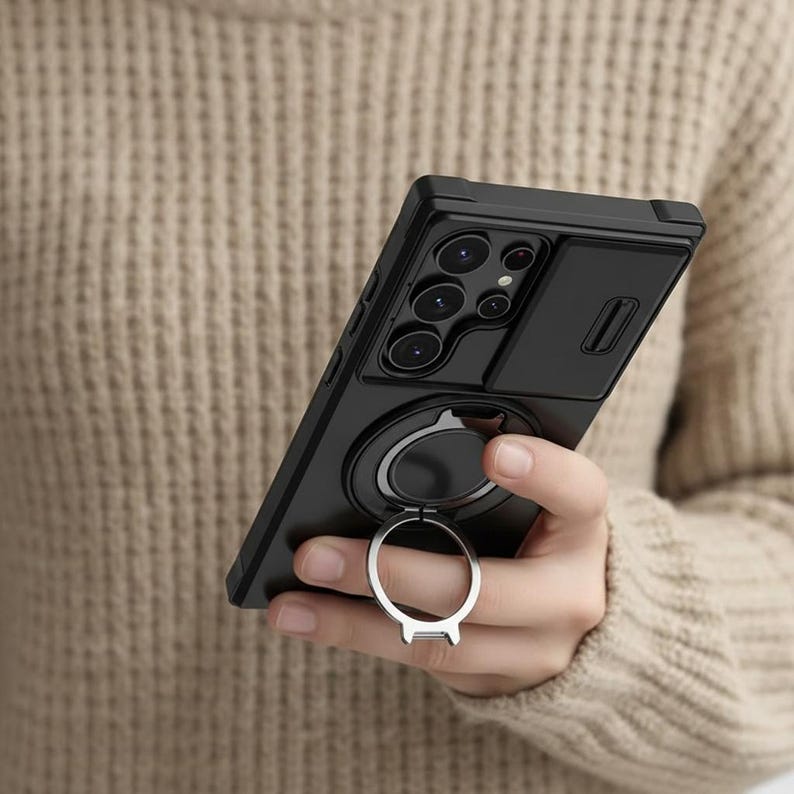 May include: Black phone case with a camera cover and a silver ring holder. The case has a matte finish and a raised edge around the camera lenses. The ring holder is attached to the back of the case and can be used as a stand.