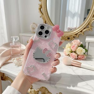 May include: Pink phone case with a Hello Kitty design. The case features a mirror, a bow, and sparkling embellishments. The camera lens is surrounded by a silver frame. The case is held in a hand.