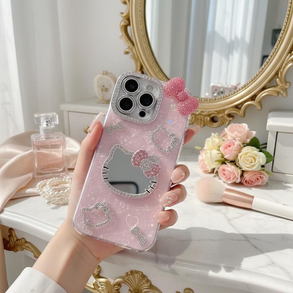 May include: Pink phone case with a Hello Kitty design. The case features a mirror, a bow, and sparkling embellishments. The camera lens is surrounded by a silver frame. The case is held in a hand.