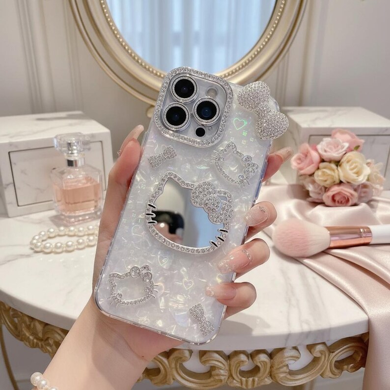 May include: A clear phone case with a white pearlescent background, adorned with sparkling rhinestones. The case features a Hello Kitty design, including a bow and character outlines. The camera lens is surrounded by a ring of rhinestones.